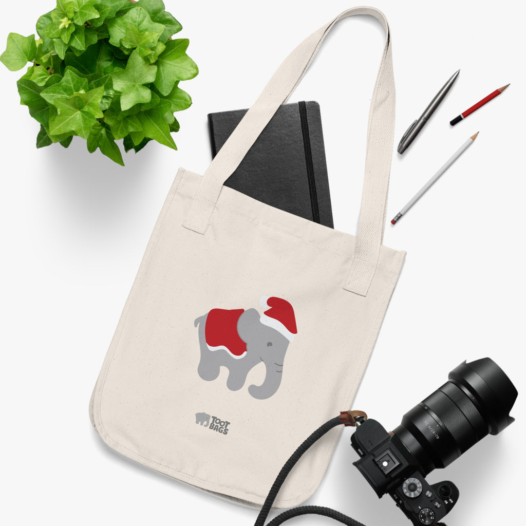 Natural canvas tote bag with printed graphics - a gray elephant wearing a red christmas hat and blanket, placed on a white surface with a plant, notebook, and camera.
