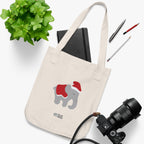 Natural canvas tote bag with printed graphics - a gray elephant wearing a red christmas hat and blanket, placed on a white surface with a plant, notebook, and camera.