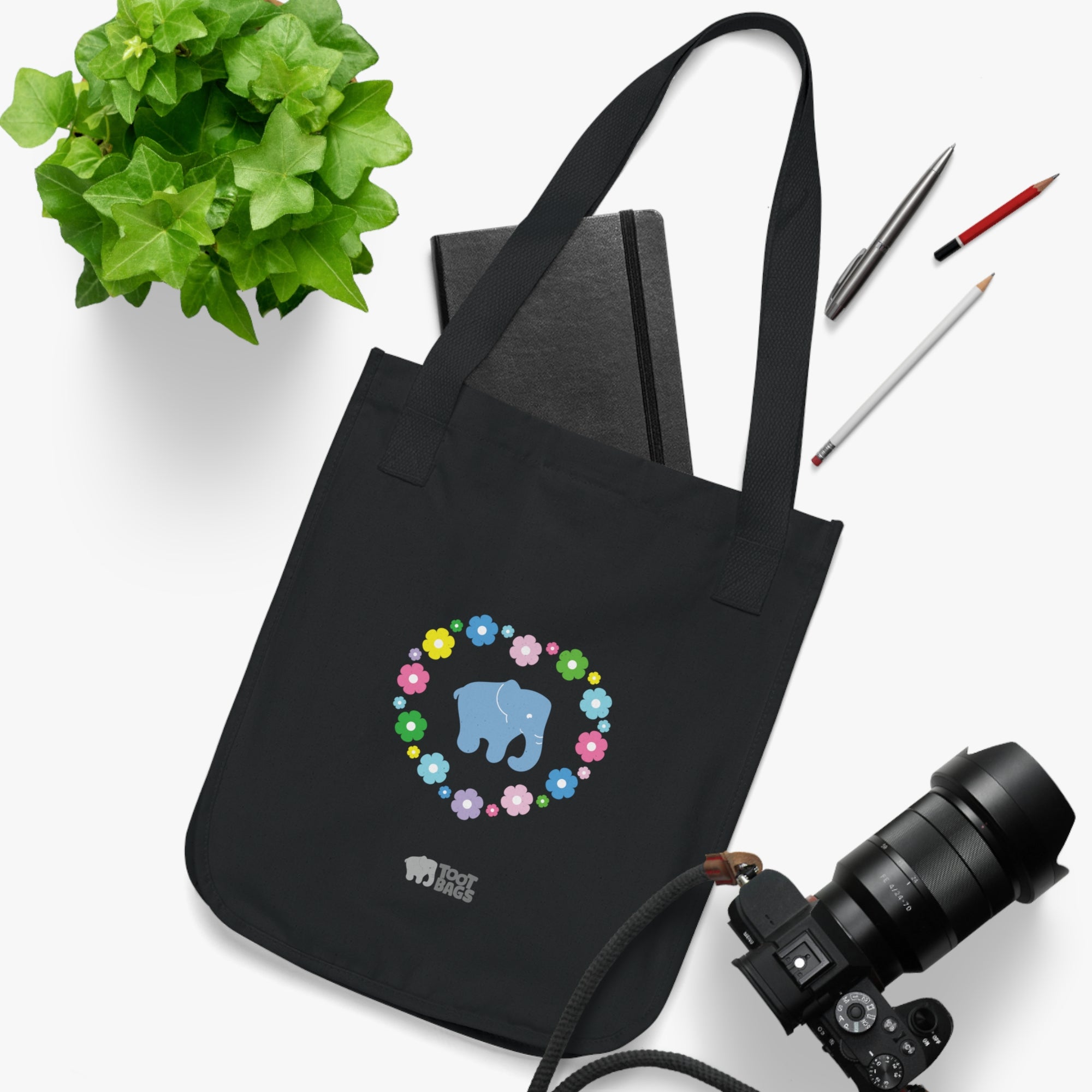 Black canvas tote bag featuring a colorful printed design; a blue elephant with floral heart shaped design placed on a white surface with stationery items, a camera and a plant.