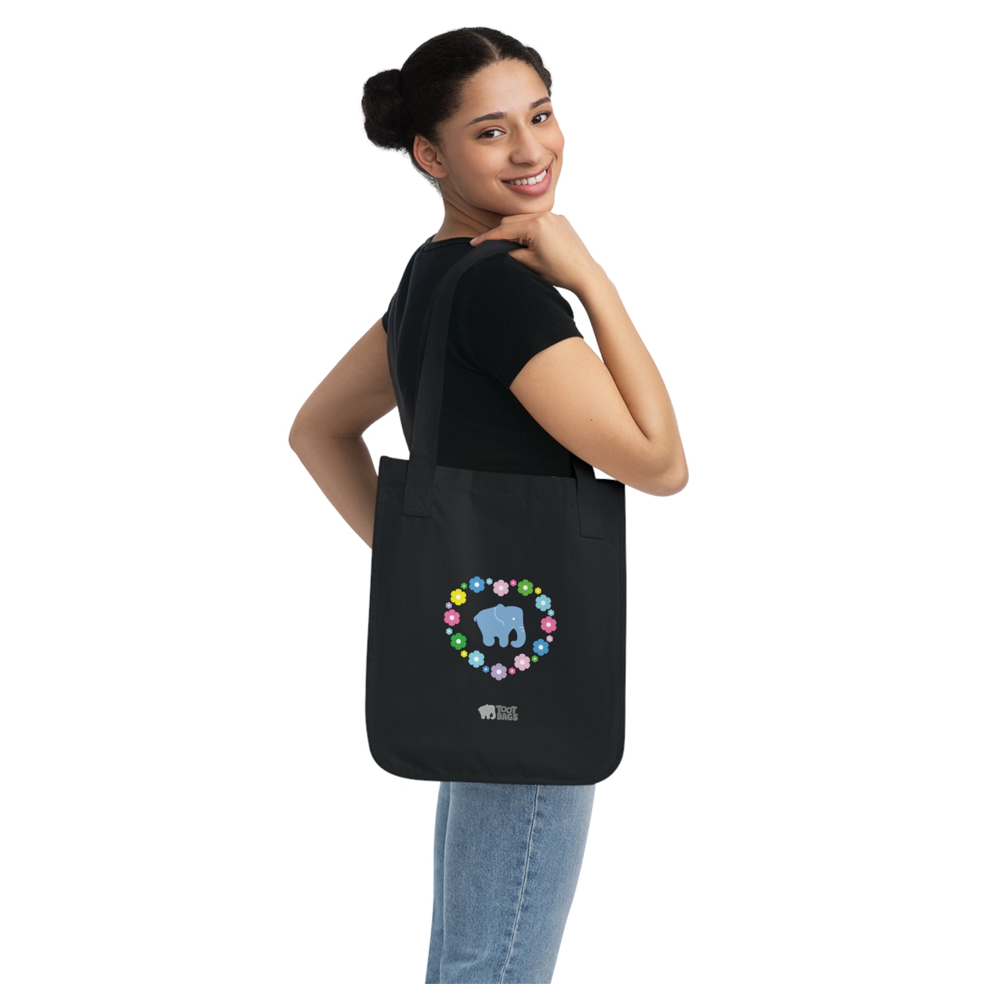 Woman carrying  a black canvas tote bag featuring a colorful printed design; a blue elephant with floral heart shaped design on a white background.  