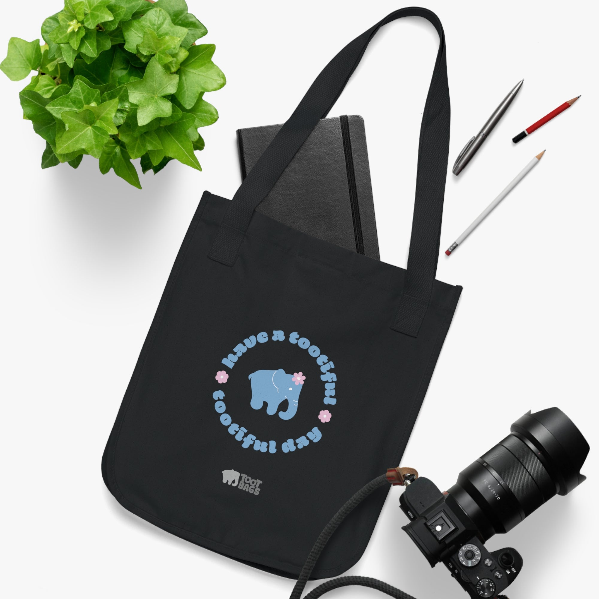 Black canvas tote bag with a printed design featuring a blue elephant and 'have a tootiful tootiful day’ text with pink floral design placed on a white surface with stationery items, a plant and a camera.