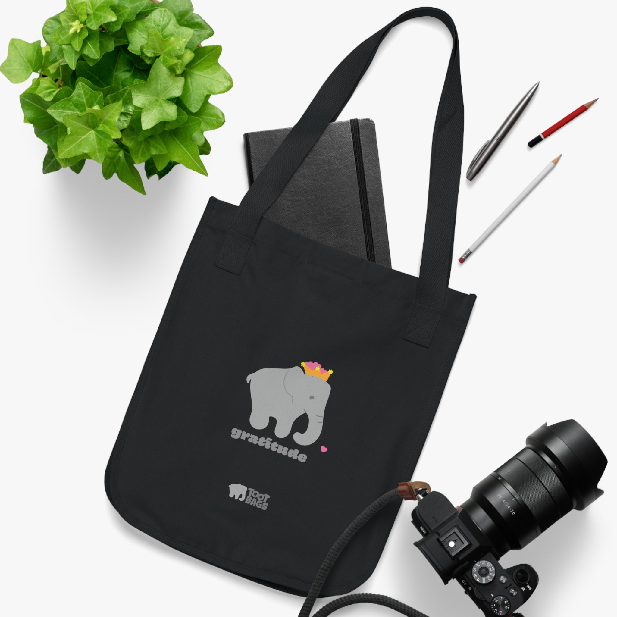 Black canvas tote bag with a printed design; a gray elephant and 'gratitude' text with colorful hearts placed on a white surface with a plant, a camera, a notebook and pencils.