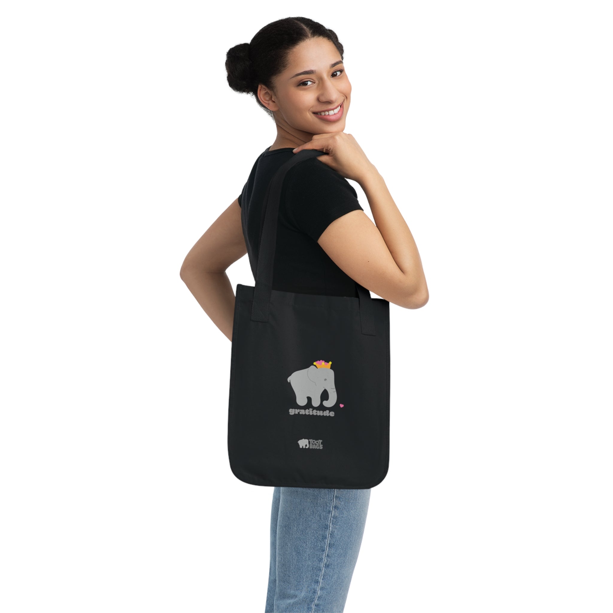 Woman carrying a black canvas tote bag featuring a printed design; a gray elephant and 'gratitude' text with colorful hearts,  on a white background.