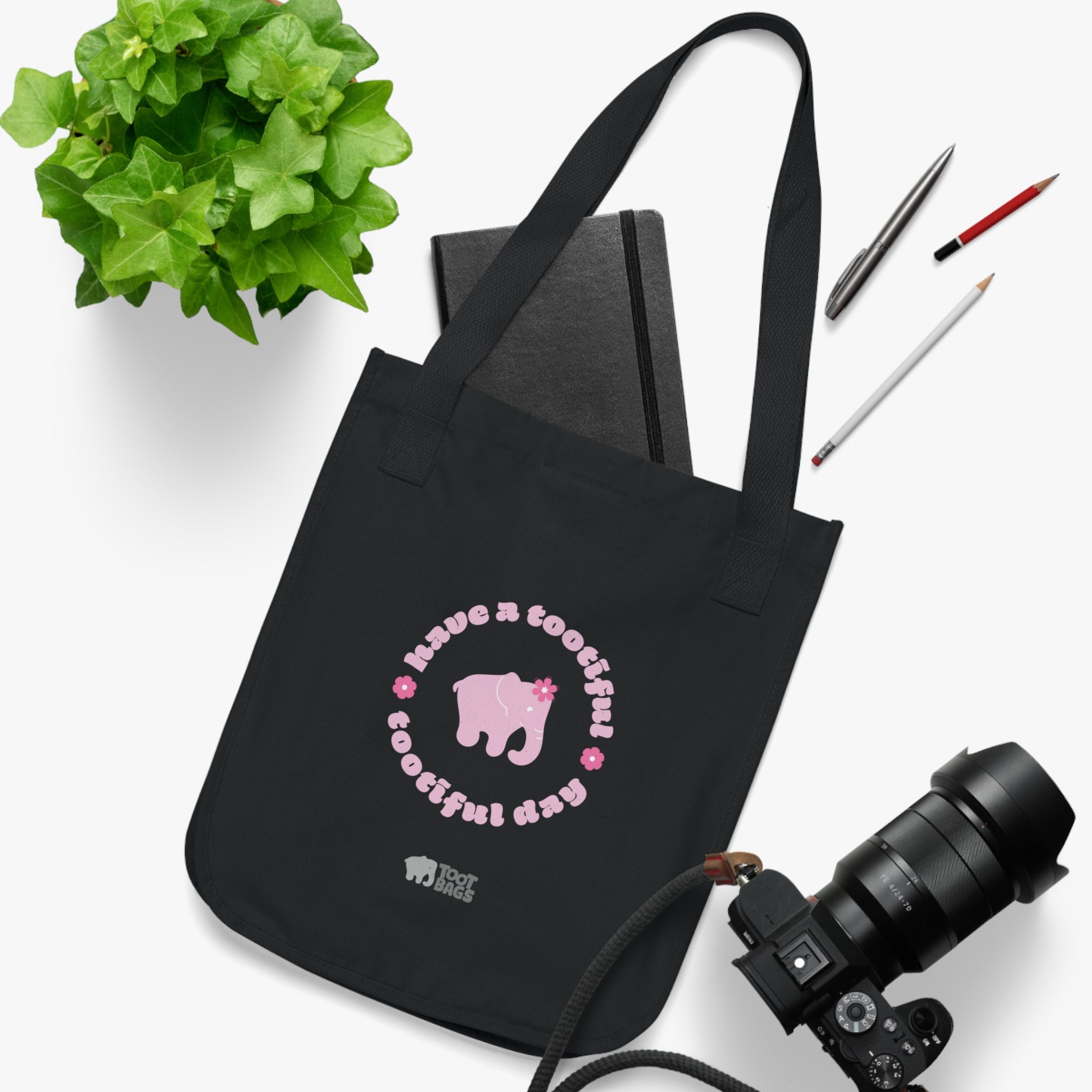 Black tote bag with a pink elephant and text with pink flowers on a white surface with stationery items.