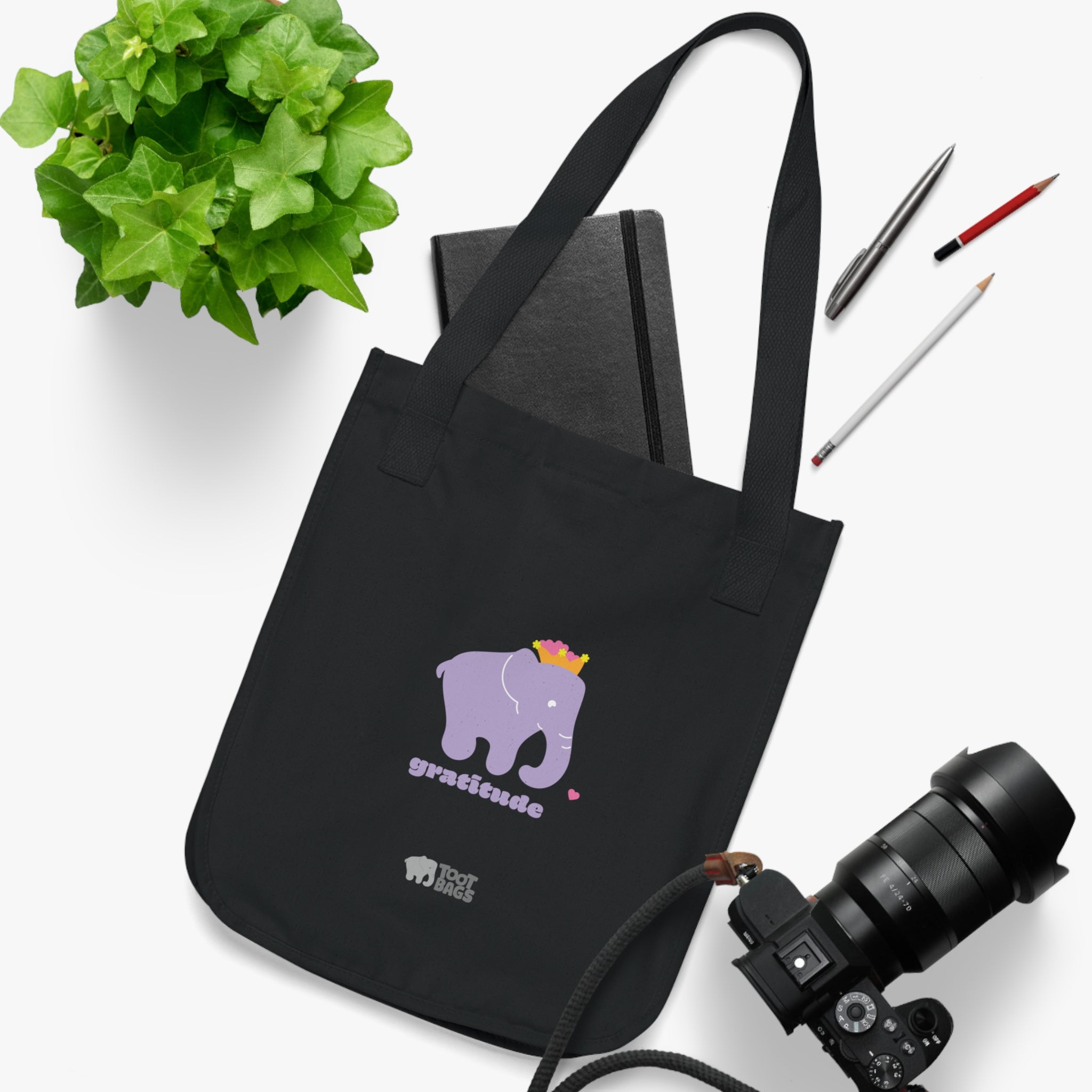 Black canvas tote bag featuring a printed design; a purple elephant, purple 'gratitude' text and colorful hearts placed on a white surface with stationery items, a plant and a camera.
