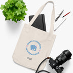 Natural beige canvas tote bag with a printed design featuring a blue elephant and 'have a tootiful tootiful day’ text with pink floral design placed on a white surface with a plant, notebook, pencils and a camera.