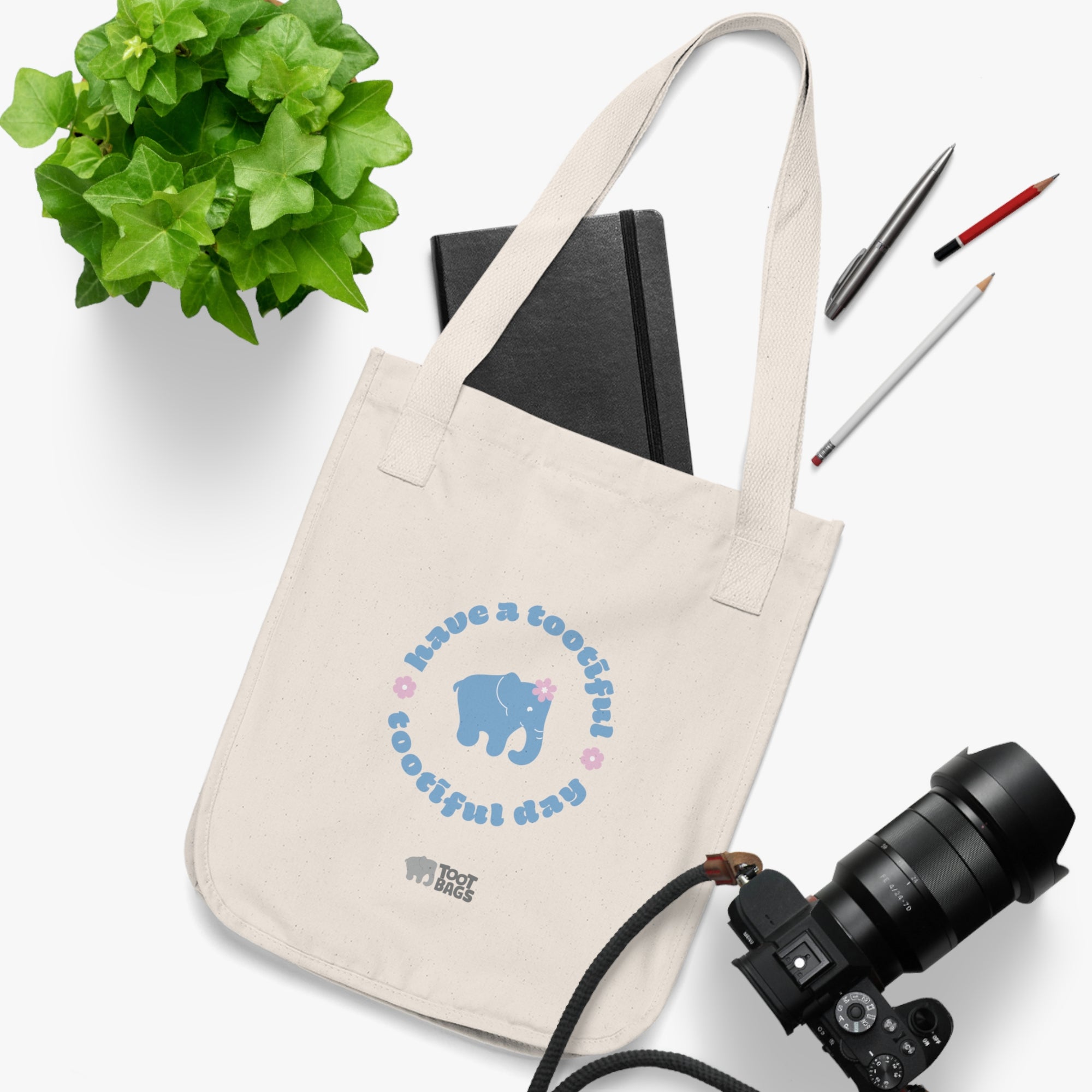 Natural beige canvas tote bag with a printed design featuring a blue elephant and 'have a tootiful tootiful day’ text with pink floral design placed on a white surface with a plant, notebook, pencils and a camera.