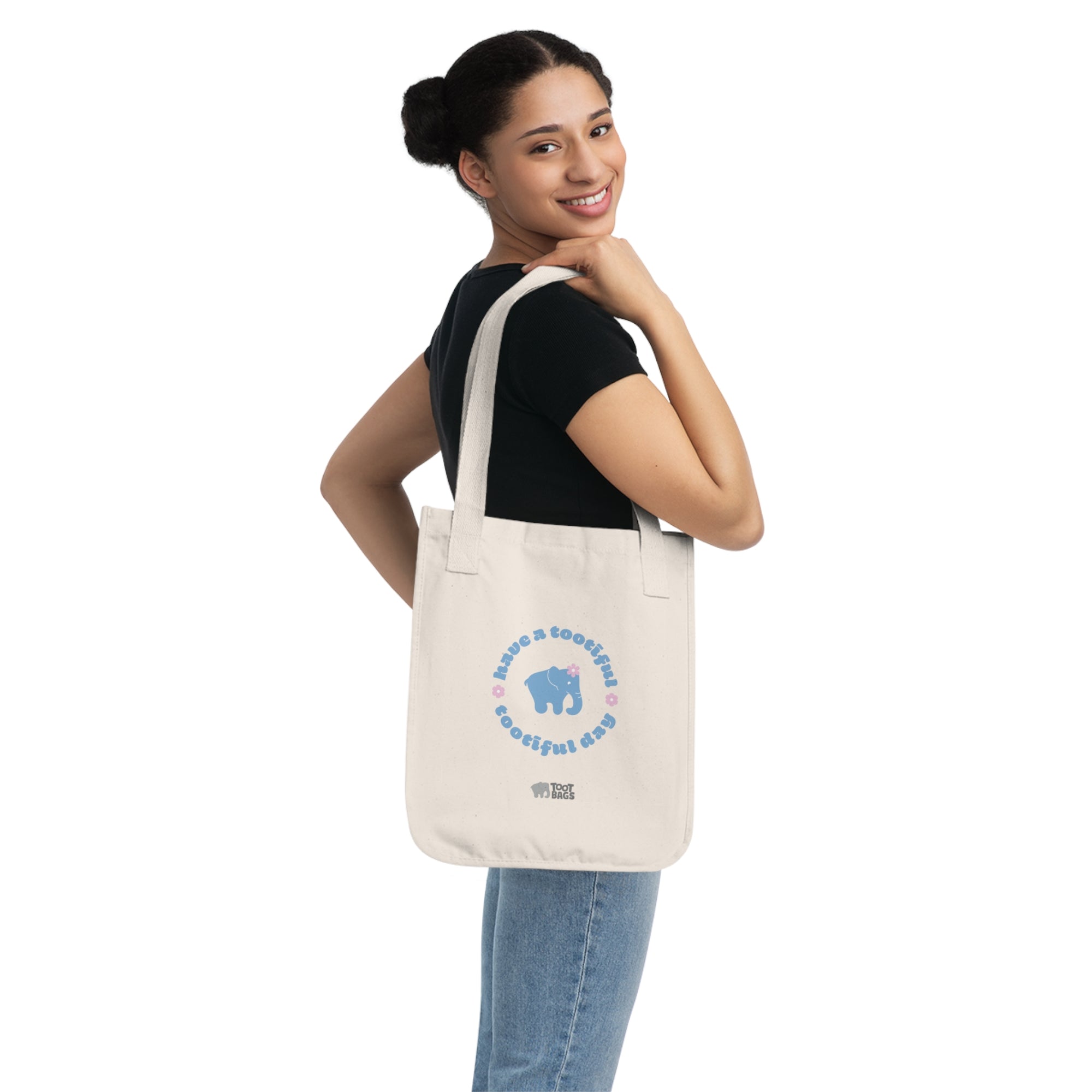 Woman holding a natural beige canvas tote bag with a printed design featuring a blue elephant and 'have a tootiful tootiful day’ text with pink floral design on a white background.
