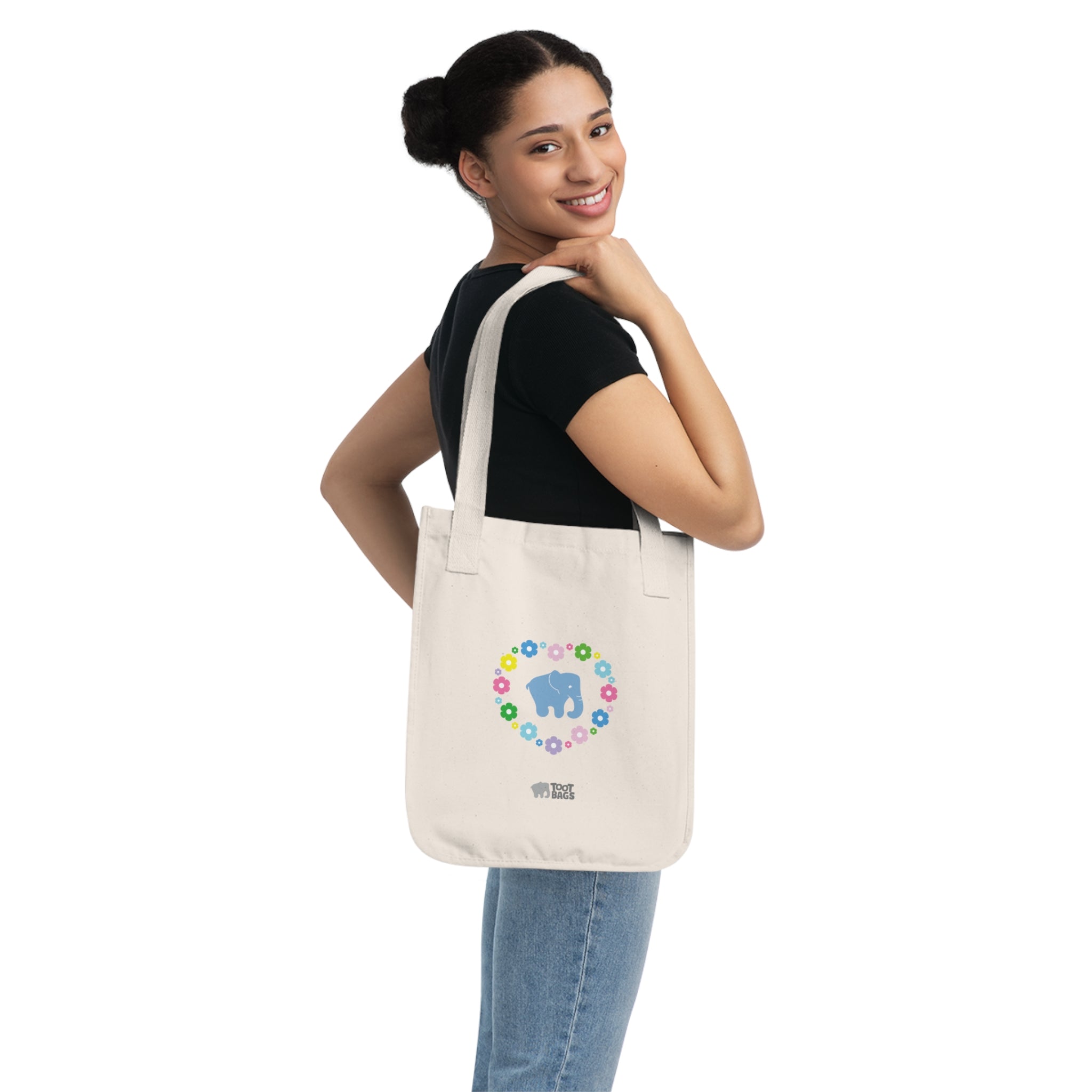 Woman holding a natural beige canvas tote bag featuring a colorful printed design; a blue elephant with floral heart shaped design on a white background.   