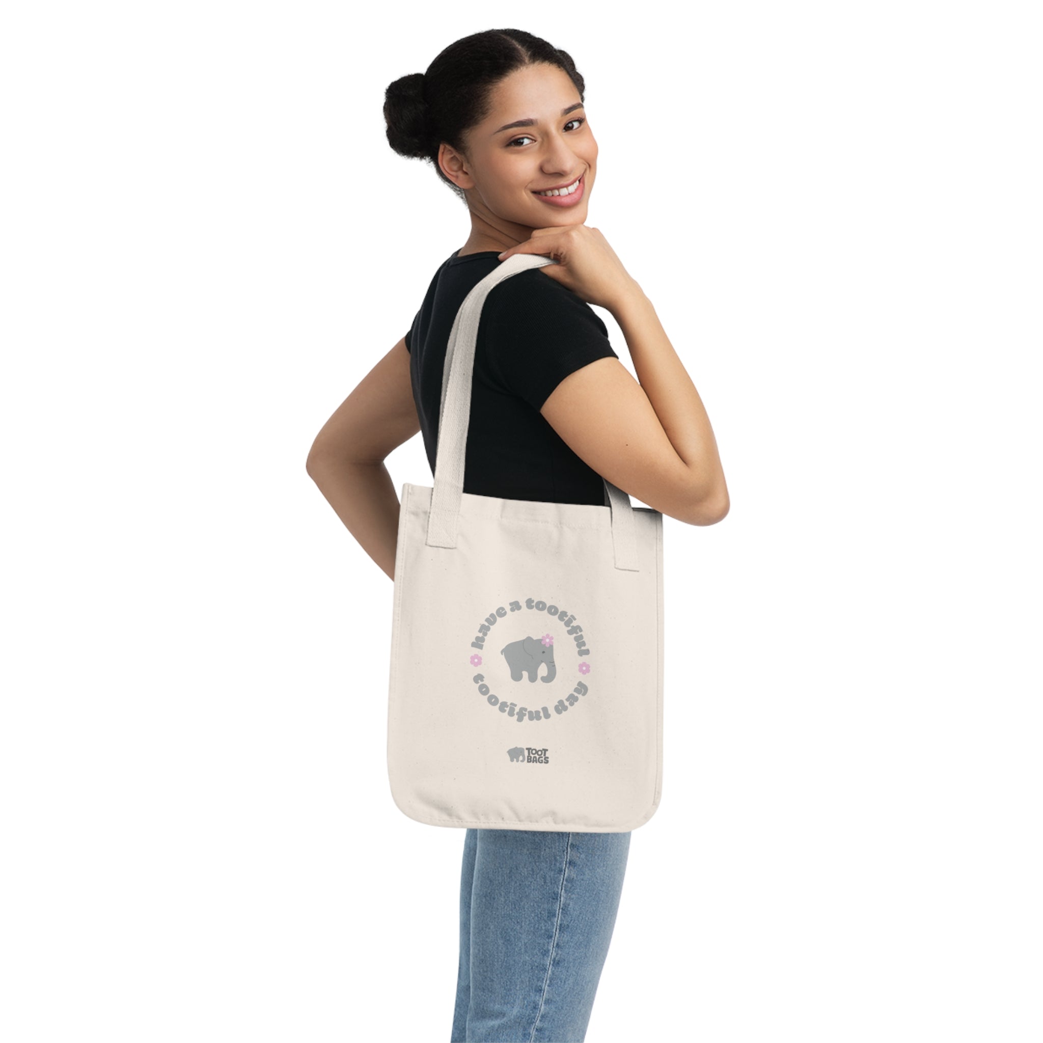 Woman holding a natural beige canvas tote bag with printed design featuring a gray elephant, gray 'have a tootiful tootiful day’ text with pink floral design on a white background.