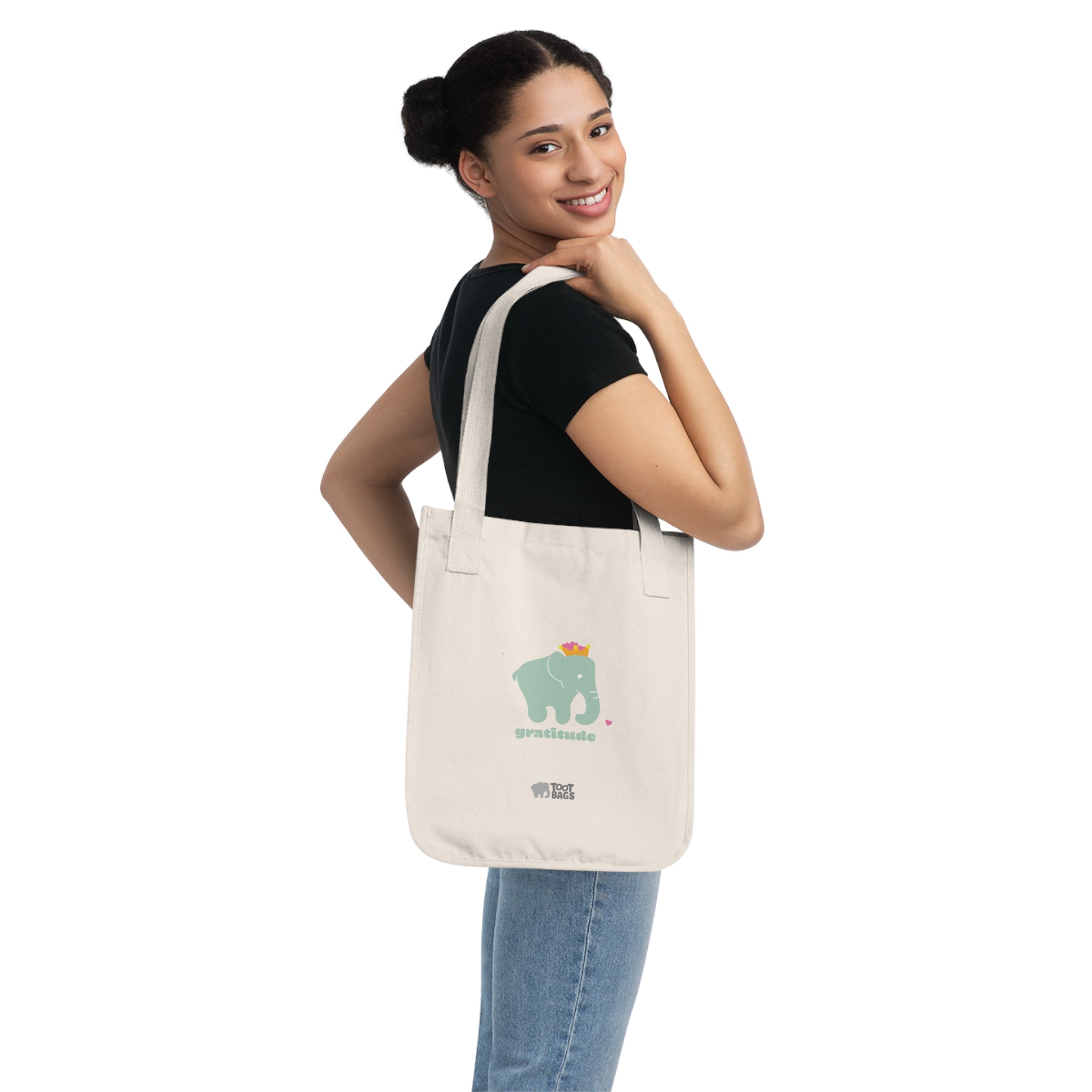 Woman carrying a beige canvas tote bag with printed design; an elephant and 'gratitude' text in matcha color with pink hearts on a white background.