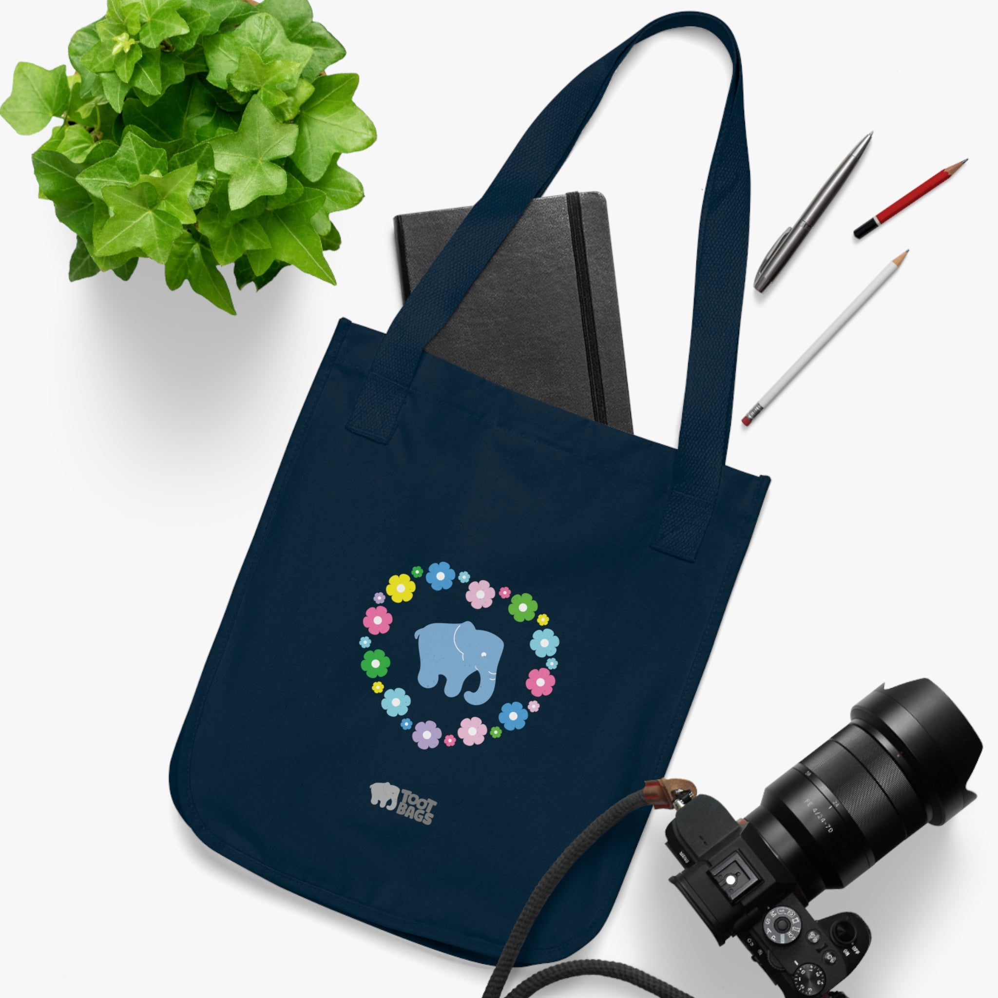 Navy blue canvas tote bag featuring a colorful printed design; a blue elephant with floral heart shaped design placed on a white surface with a plant, camera, notebook and pencils.