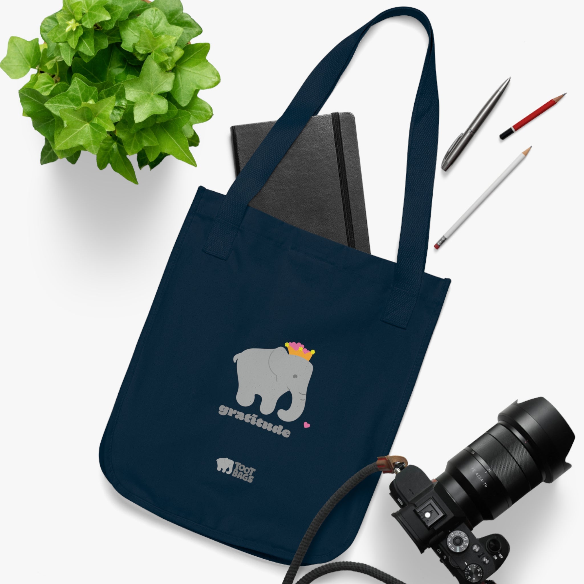 Navy canvas tote bag with a printed design; a gray elephant and 'gratitude' text with colorful hearts placed on a white surface with a plant, a camera, a notebook and pencils.