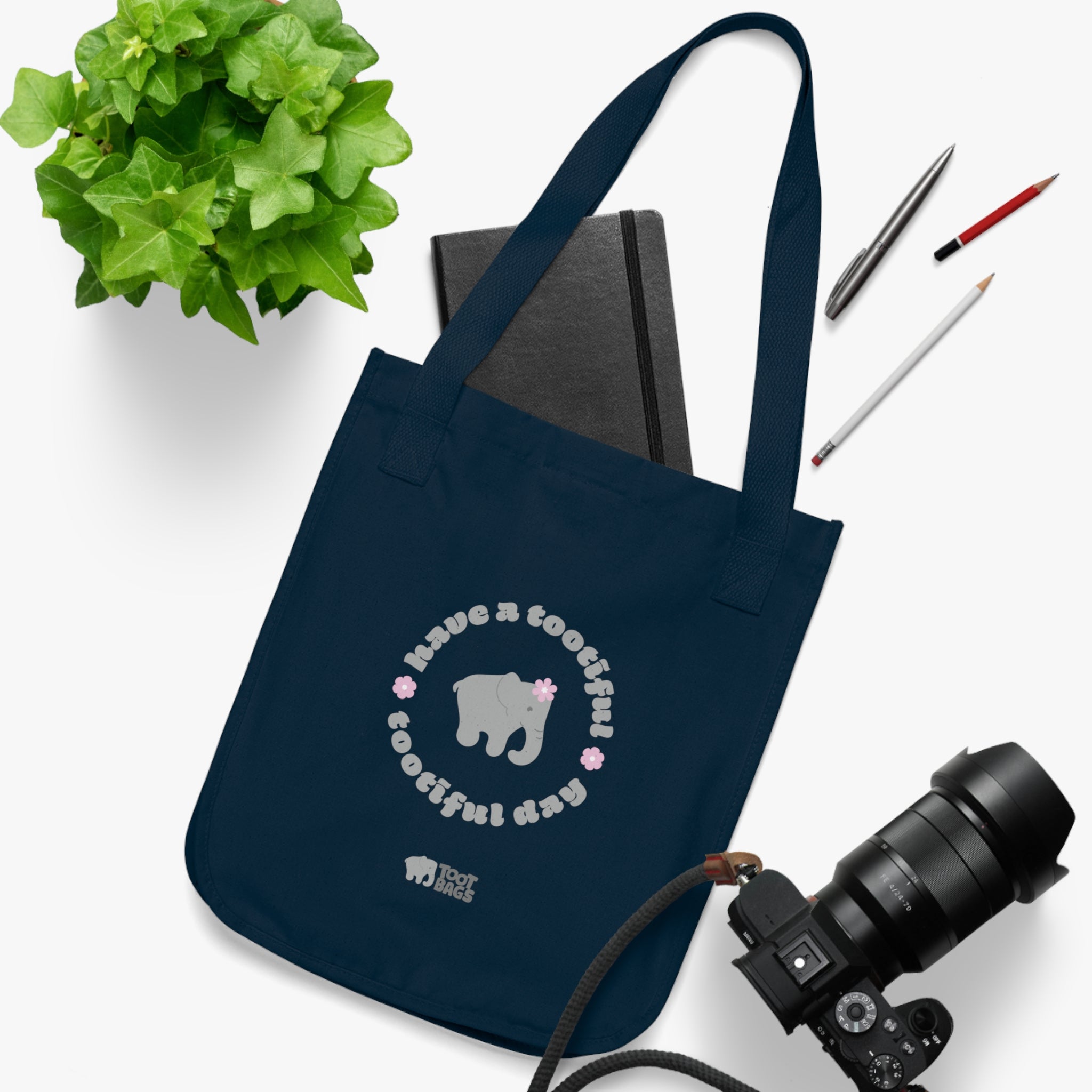 Navy blue canvas tote bag with printed design featuring a gray elephant, gray 'have a tootiful tootiful day’ text with pink floral design on a white surface with stationery items, a plant and a camera.