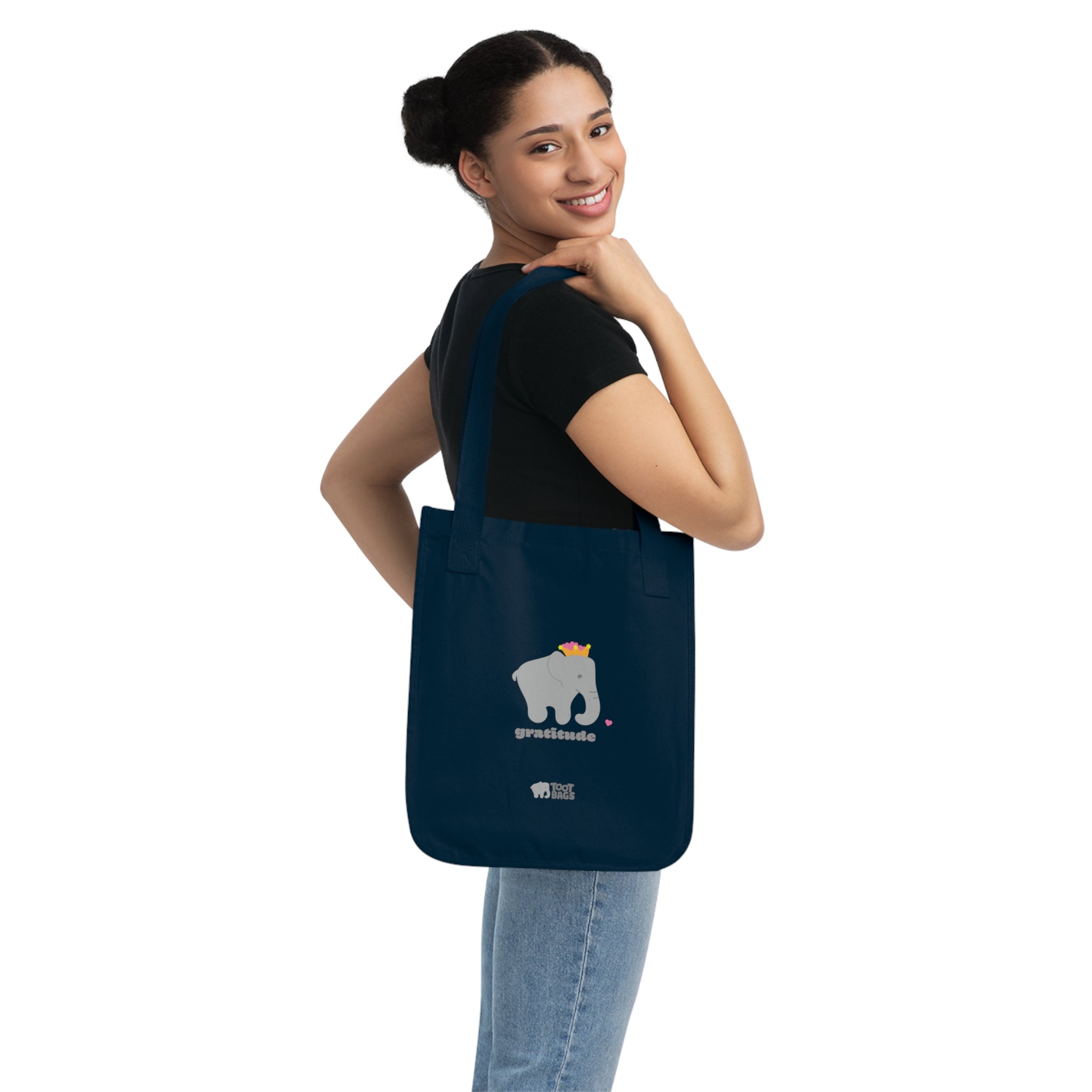 Woman carrying a navy blue canvas tote bag featuring a printed design; a gray elephant and gratitude text with colorful hearts, on a white background.