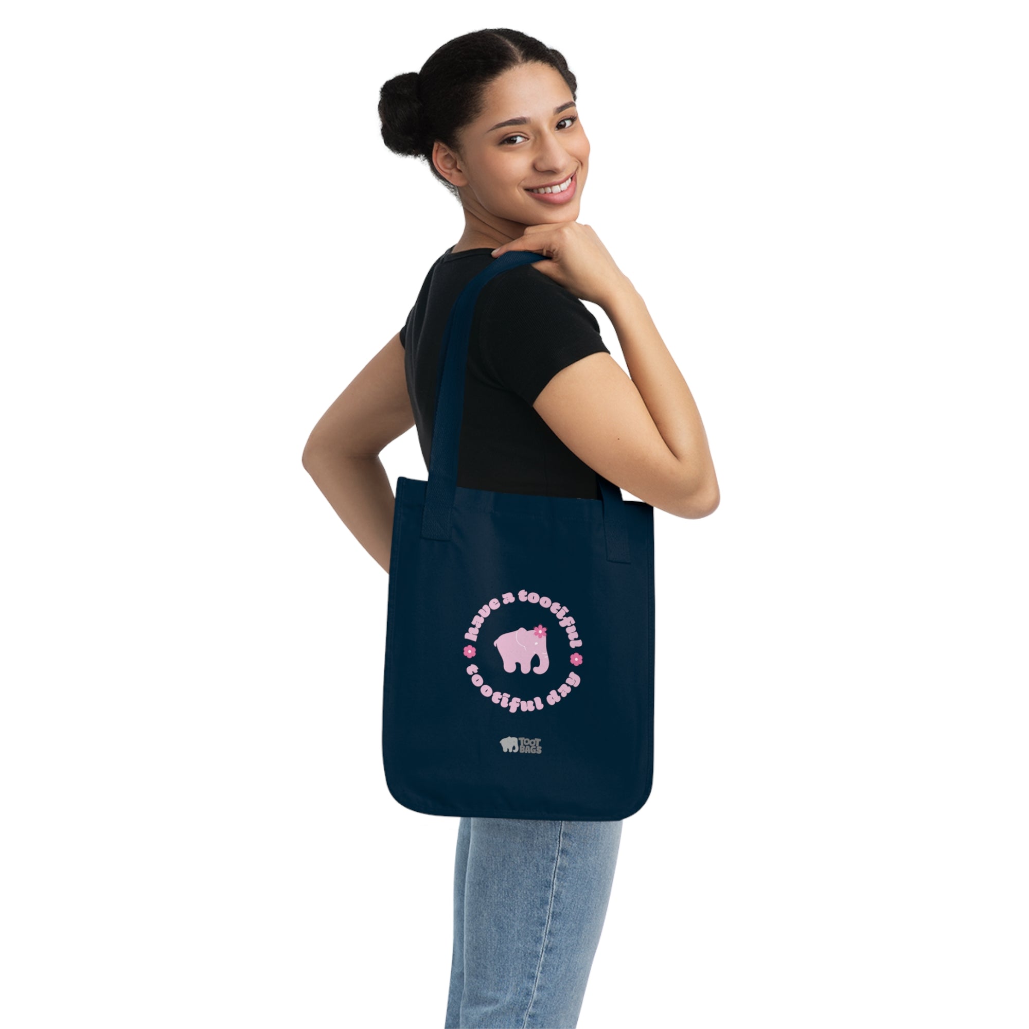 Woman carrying a navy blue canvas tote bag with printed design featuring a pink elephant, pink 'have a tootiful tootiful day’ text with colorful pink flowers on a white background.
