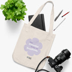 Natural canvas tote bag with a printed design featuring a purple flower and ‘the peaceful state of gratitude’ text placed on a white surface with stationery items, a plant and a camera.