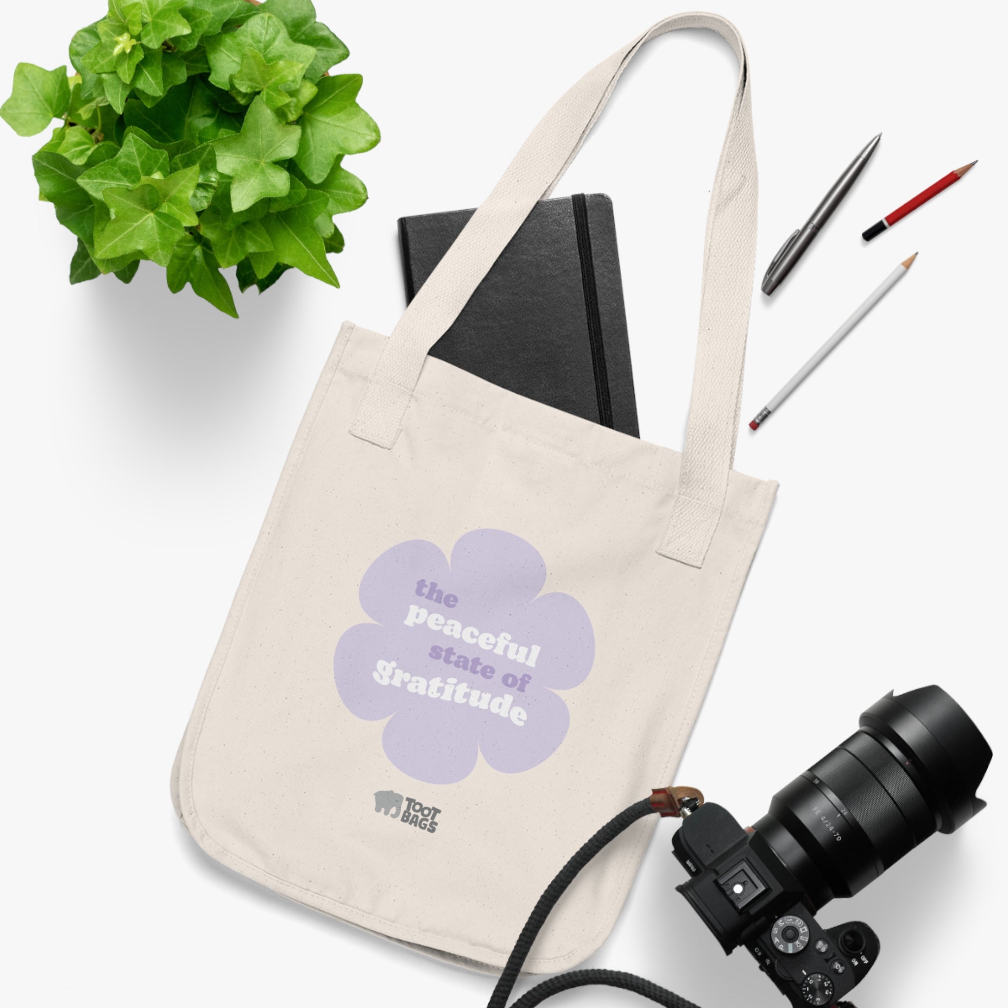 Natural canvas tote bag with a printed design featuring a purple flower and ‘the peaceful state of gratitude’ text placed on a white surface with stationery items, a plant and a camera.