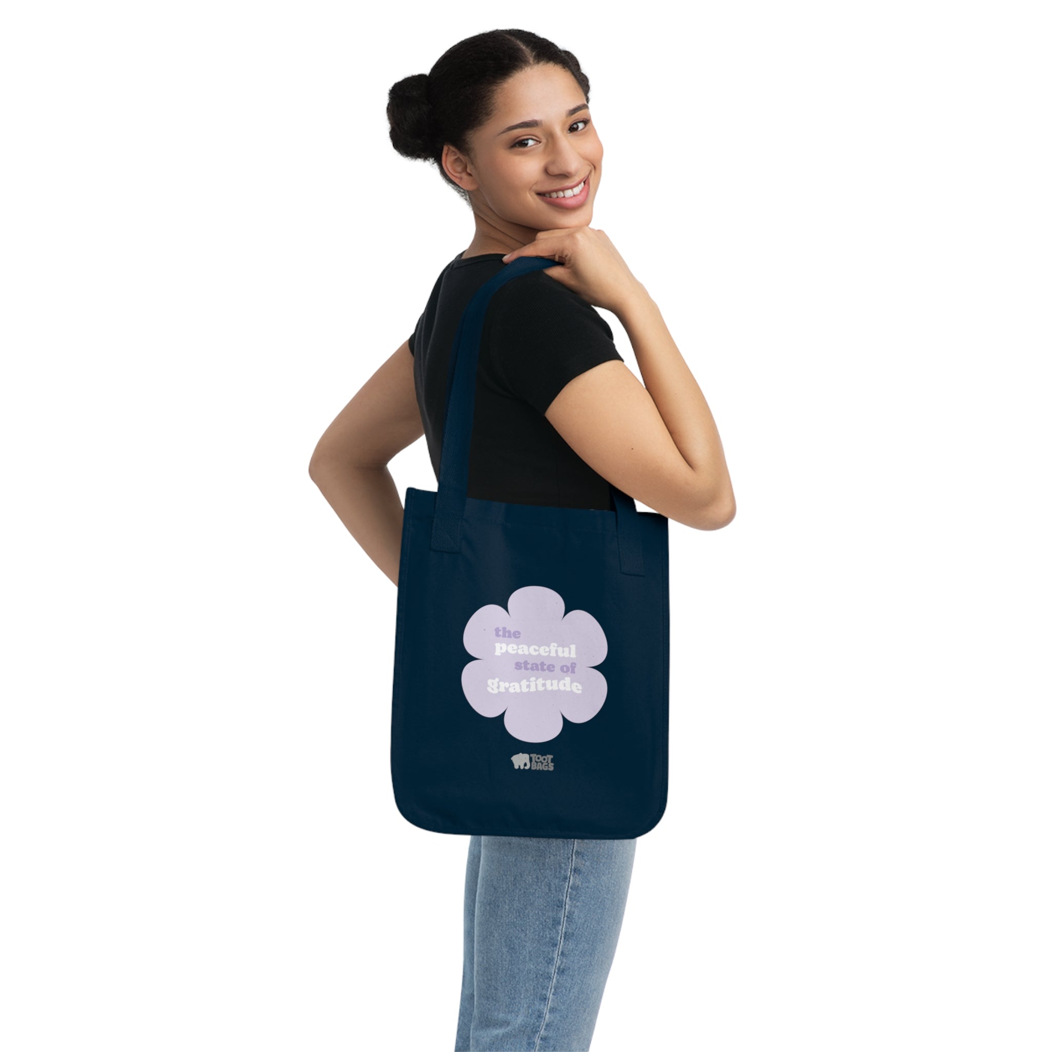 Woman holding a navy blue canvas tote bag with a printed design featuring a purple flower and ‘the peaceful state of gratitude’ text design in purple and white on a white background.