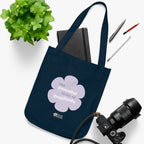 Navy blue canvas tote bag with a printed design featuring a purple flower and ‘the peaceful state of gratitude’ text  placed on a white surface with stationery items, a plant and a camera.