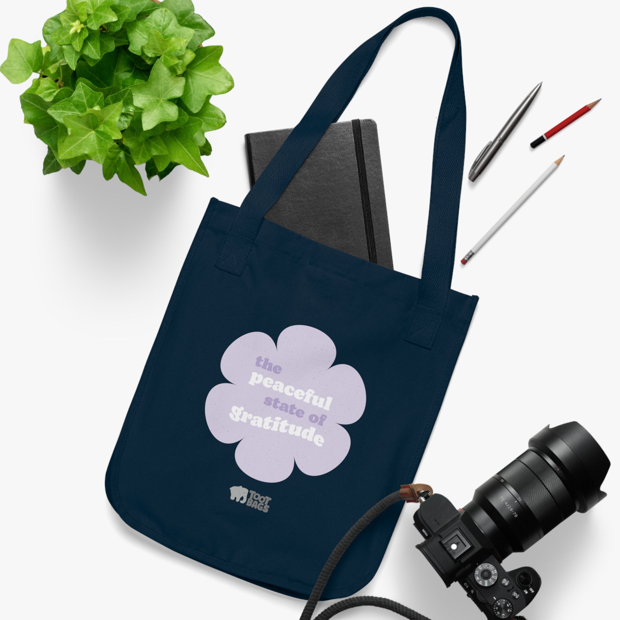 Navy blue canvas tote bag with a printed design featuring a purple flower and ‘the peaceful state of gratitude’ text  placed on a white surface with stationery items, a plant and a camera.