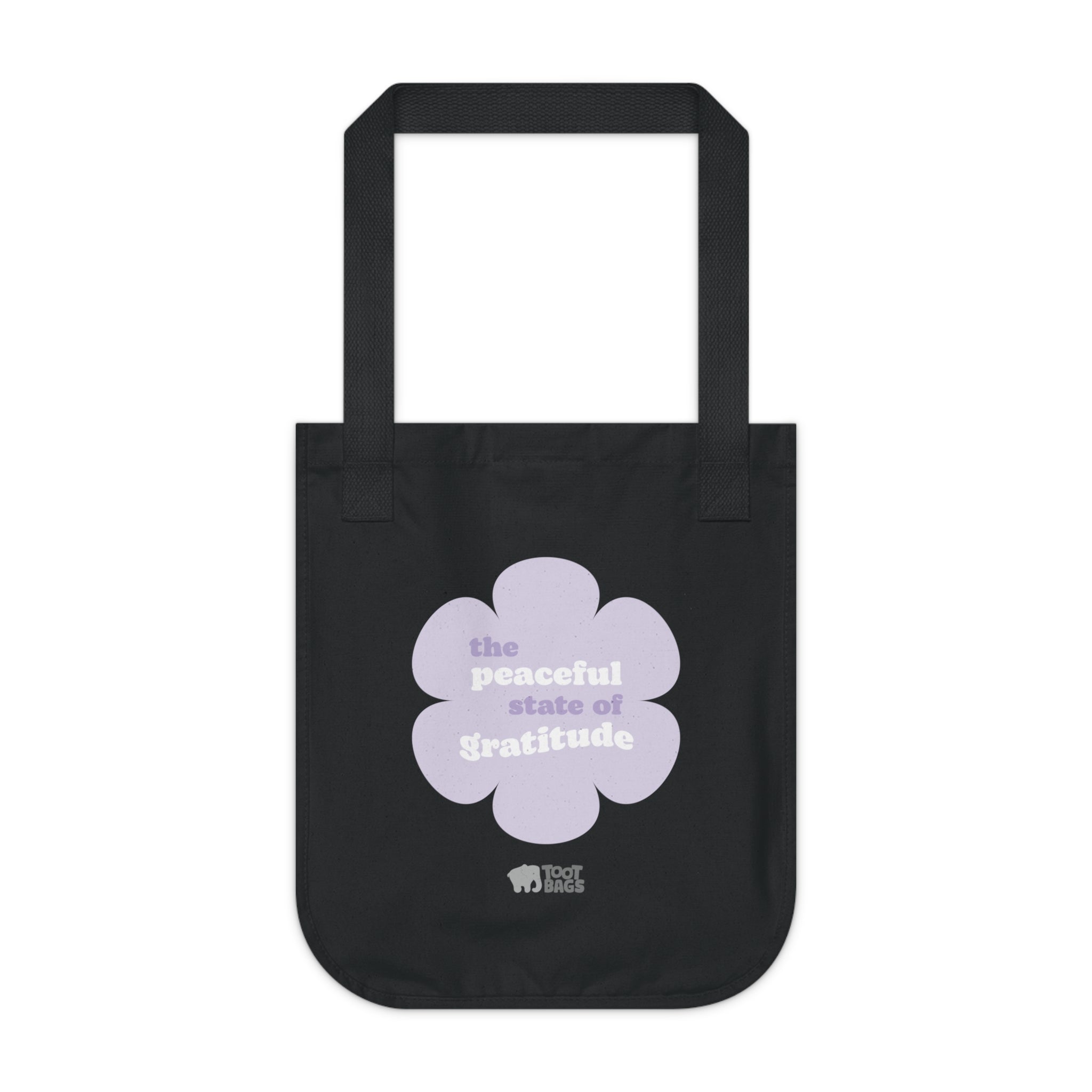 Black canvas tote bag with a printed design featuring a purple flower and ‘the peaceful state of gratitude’ text design in purple and white placed on a white background.