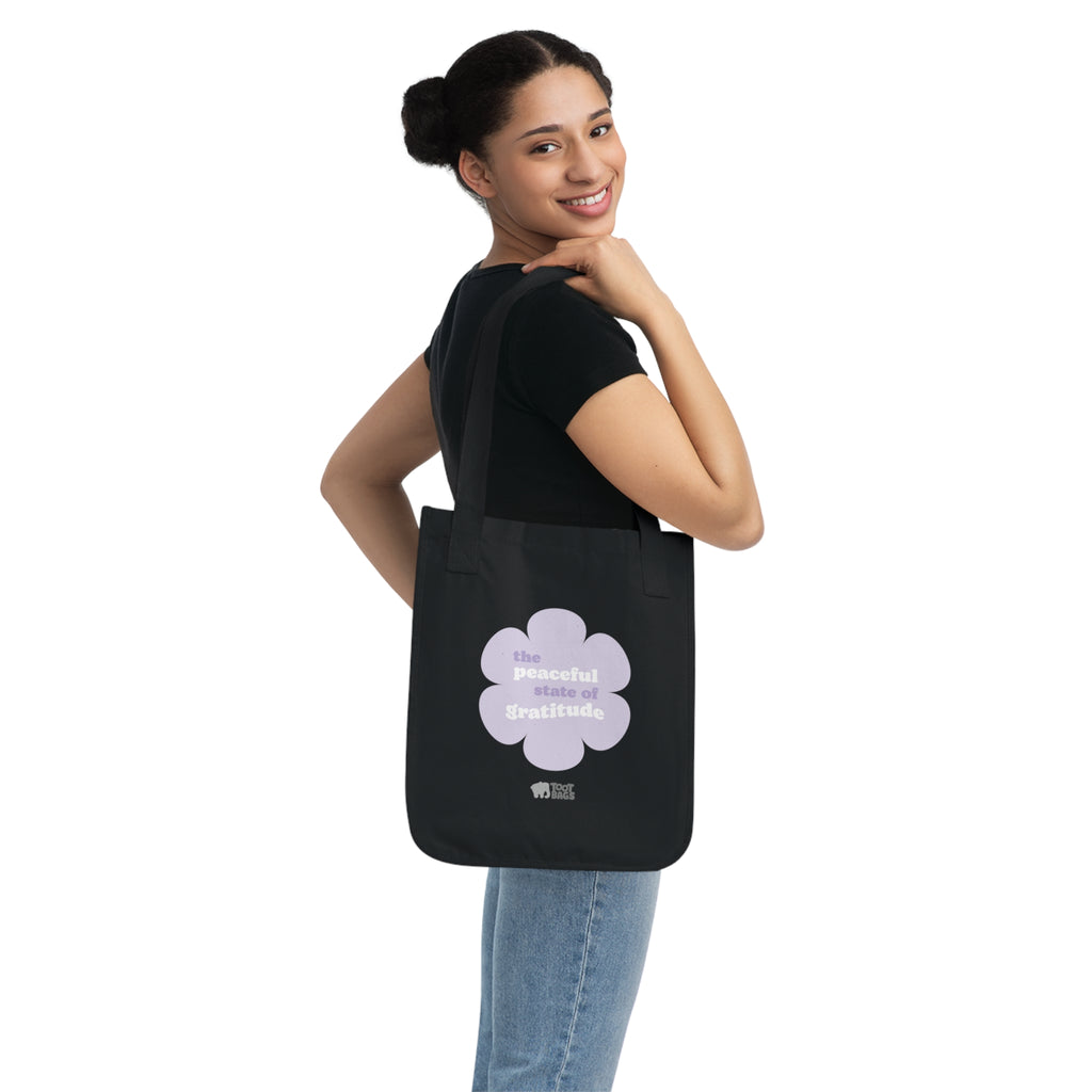 Woman holding a black canvas tote bag with a printed design featuring a purple flower and ‘the peaceful state of gratitude’ text design in purple and white on a white background.