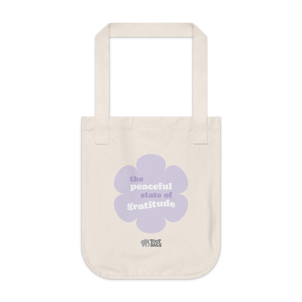 Natural beige canvas tote bag with a printed design featuring a purple flower and ‘the peaceful state of gratitude’ text design in purple and white placed on a white background.