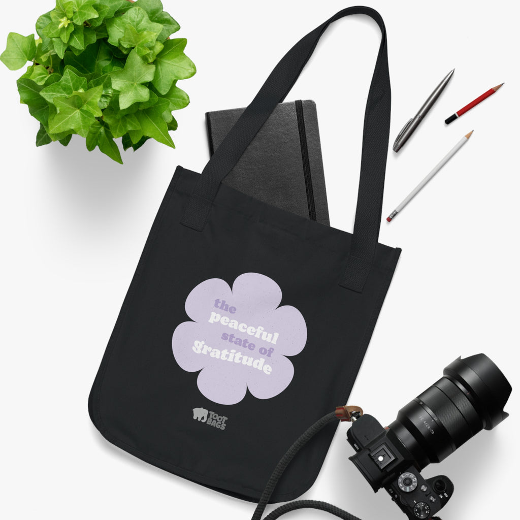 Black canvas tote bag with a printed design featuring a purple flower and ‘the peaceful state of gratitude’ text placed on a white surface with a plant, notebook, and camera.