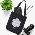 Black canvas tote bag with a printed design featuring a purple flower and ‘the peaceful state of gratitude’ text placed on a white surface with a plant, notebook, and camera.