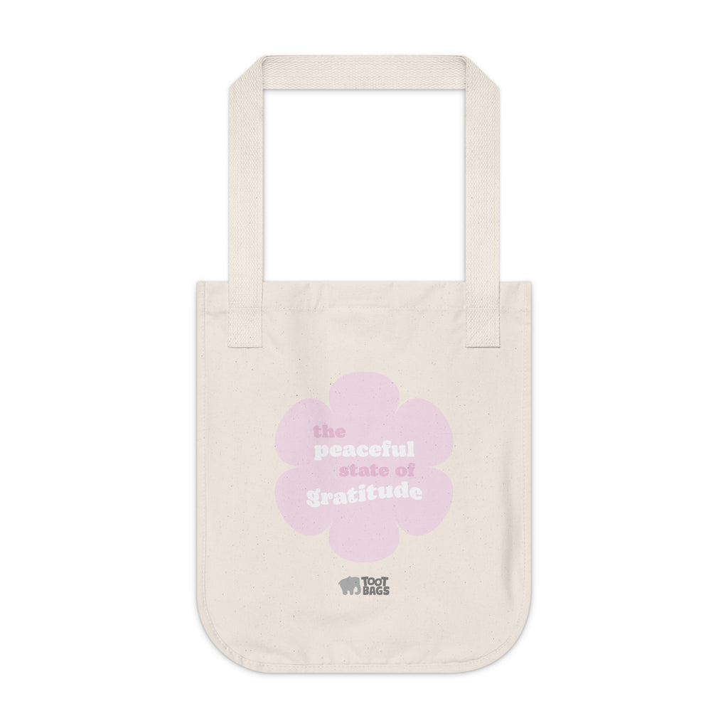 Natural beige canvas tote bag with a printed design featuring a pink flower and ‘the peaceful state of gratitude’ text design placed on a white background.
