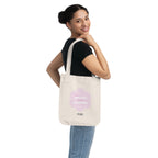 Woman holding a natural beige canvas tote bag with a printed design featuring a pink flower and ‘the peaceful state of gratitude’ text design on a white background.
