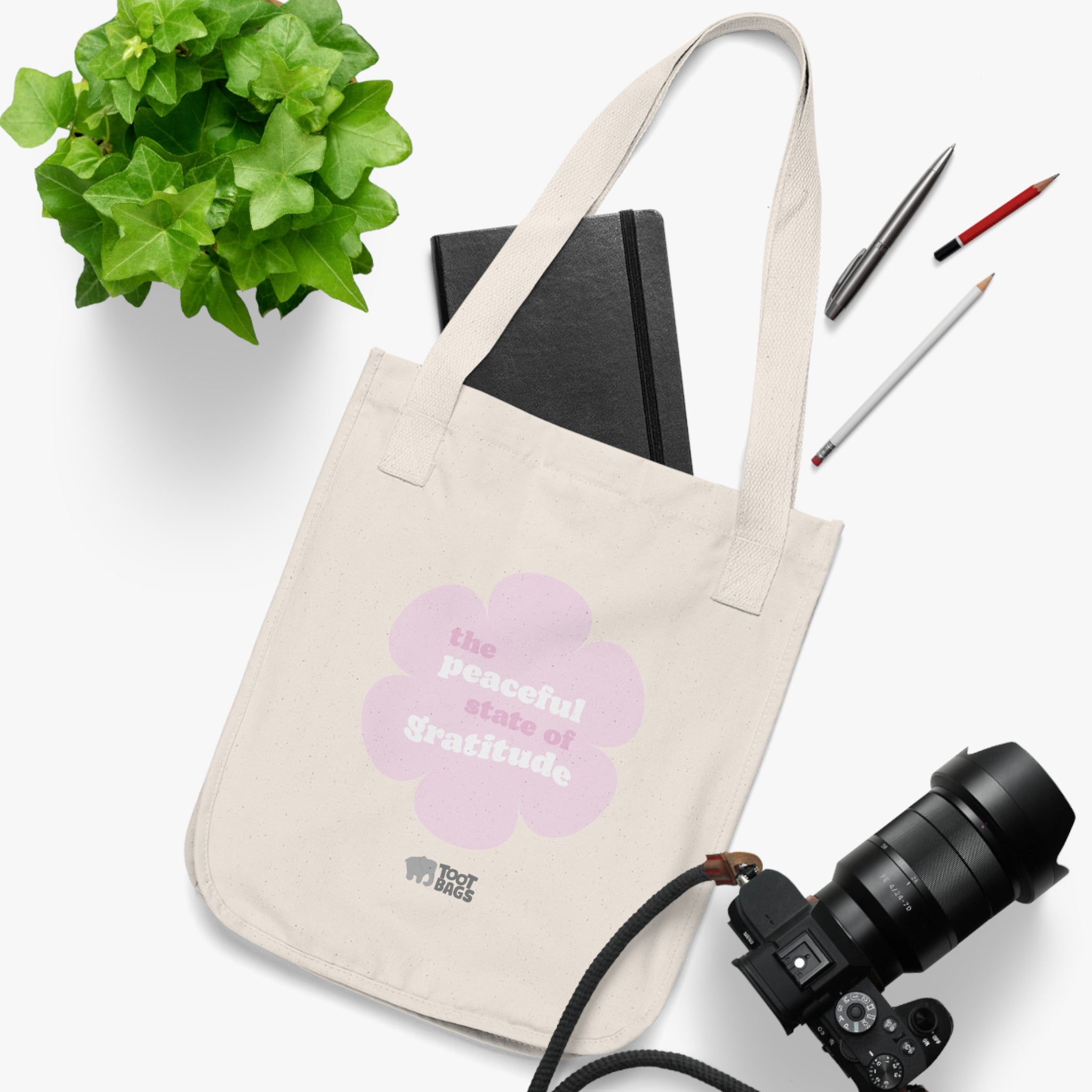 Natural beige canvas tote bag with a printed design featuring a pink flower and ‘the peaceful state of gratitude’ text placed on a white surface with a plant, notebook, and camera.