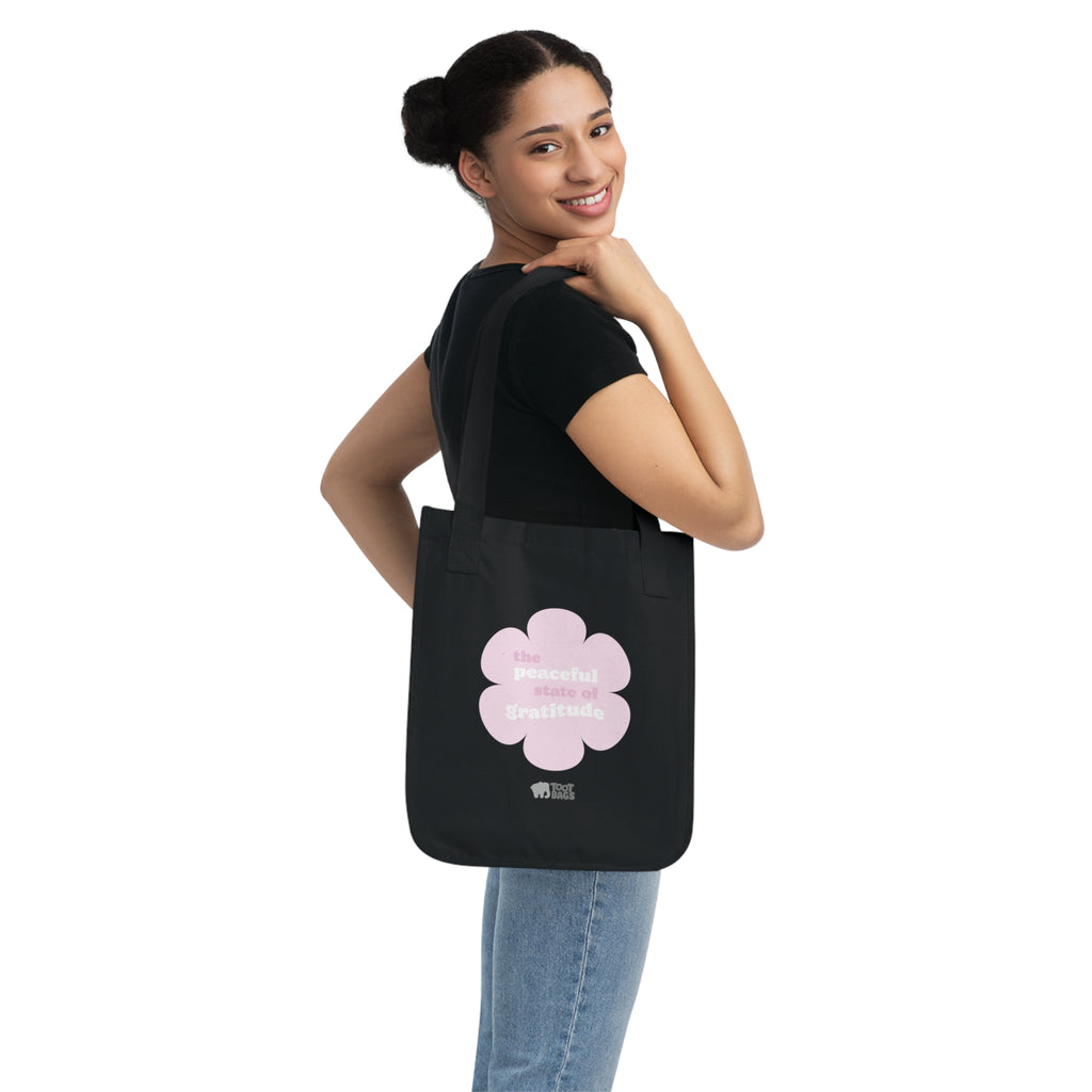 Woman holding a black canvas tote bag with a printed design featuring a pink flower and ‘the peaceful state of gratitude’ text design on a white background.
