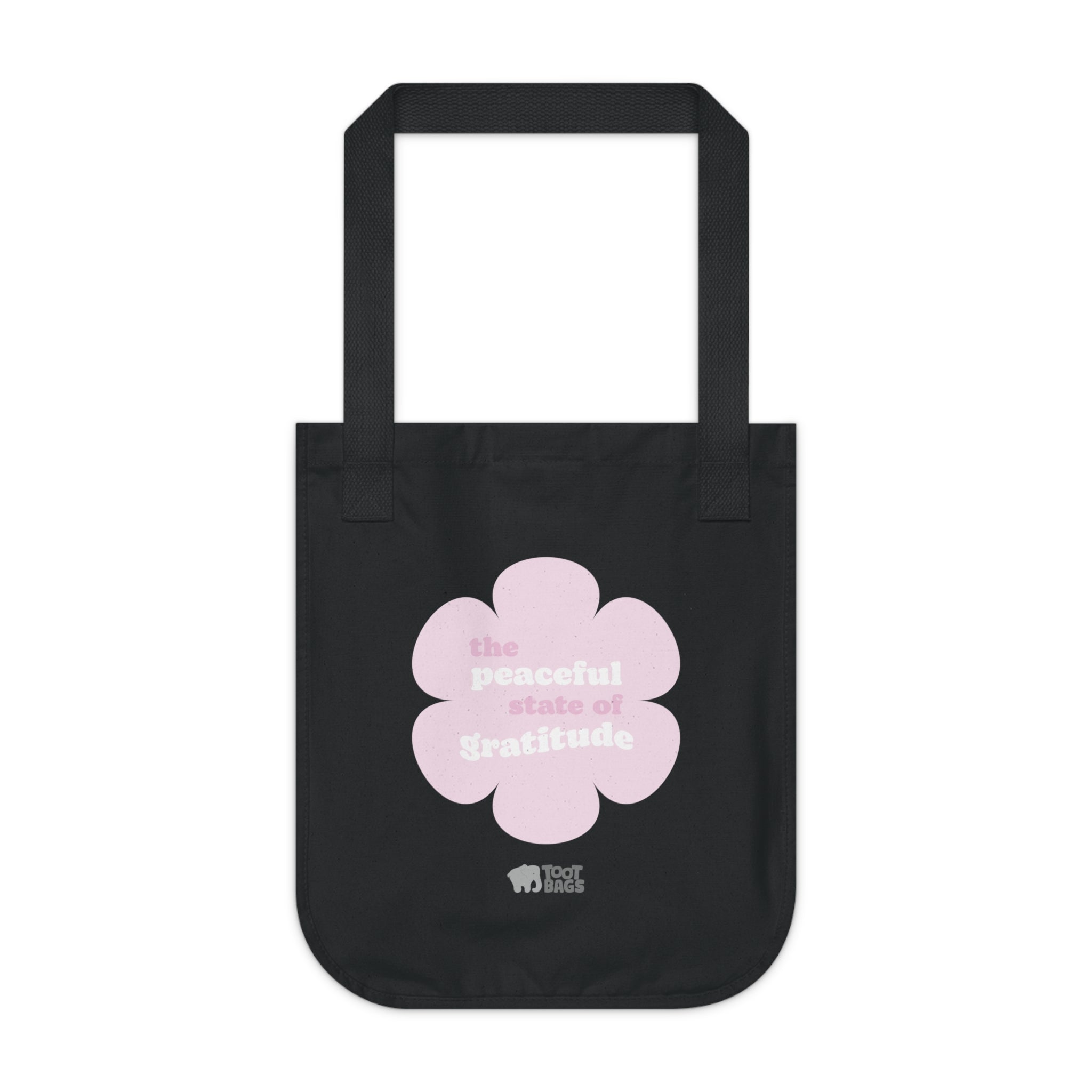 Black canvas tote bag with a printed design featuring a pink flower and ‘the peaceful state of gratitude’ text design placed on a white background.