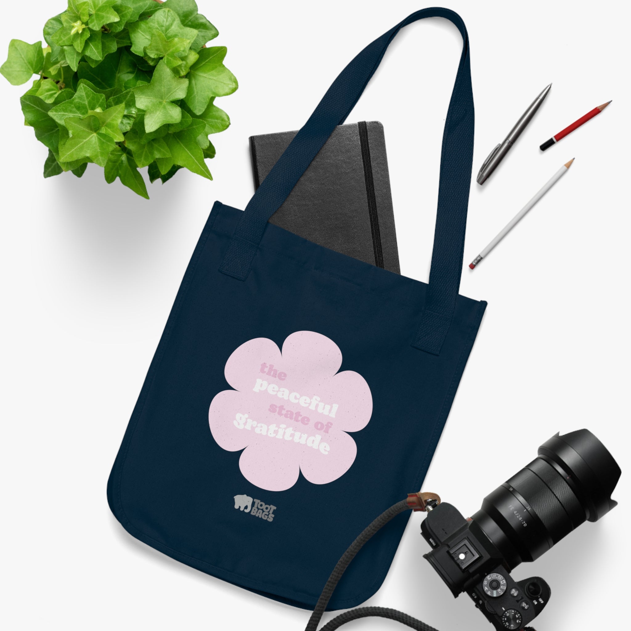Navy blue canvas tote bag with a printed design featuring a pink flower and ‘the peaceful state of gratitude’ text placed on a white surface with a plant, notebook, and camera.