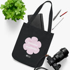 Black canvas tote bag with a printed design featuring a pink flower and ‘the peaceful state of gratitude’ text placed on a white surface with a plant, notebook, and camera.