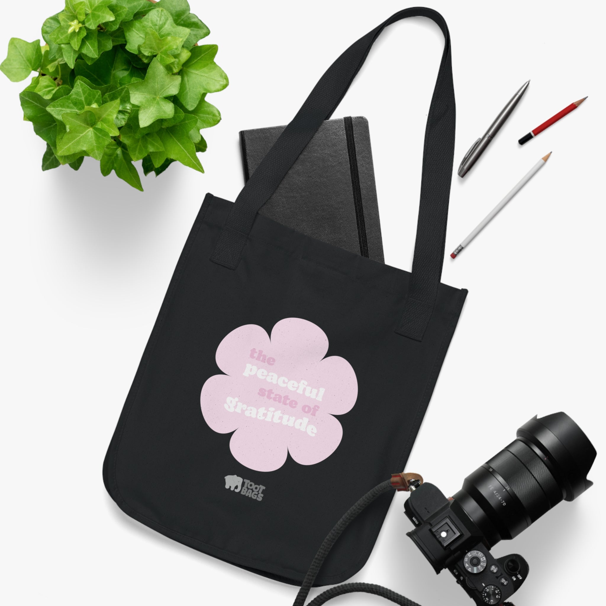 Black canvas tote bag with a printed design featuring a pink flower and ‘the peaceful state of gratitude’ text placed on a white surface with a plant, notebook, and camera.