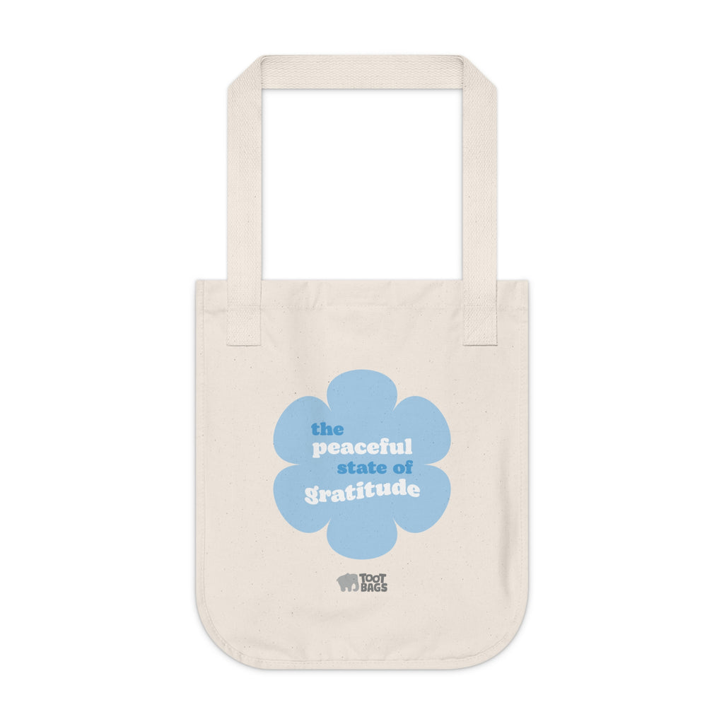 Natural beige canvas tote bag with a printed design featuring a blue flower and ‘the peaceful state of gratitude’ text design on a white background.