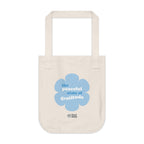 Natural beige canvas tote bag with a printed design featuring a blue flower and ‘the peaceful state of gratitude’ text design on a white background.