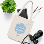 Natural beige canvas tote bag with a printed design featuring a blue flower and ‘the grateful state of gratitude’ text design placed on a white surface with stationery items, a plant and a camera.