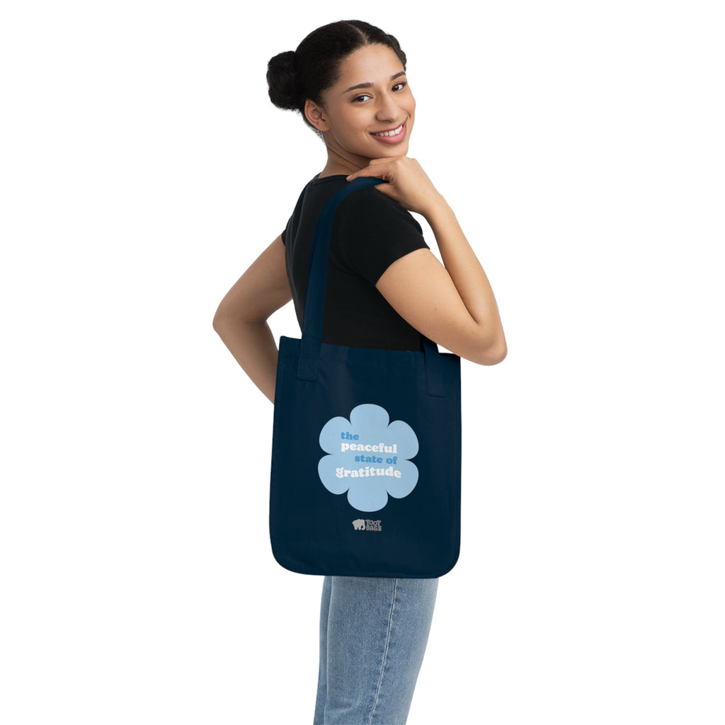 Woman holding a navy blue canvas tote bag with a printed design featuring a blue flower and ‘the peaceful state of gratitude’ text design on a white background.