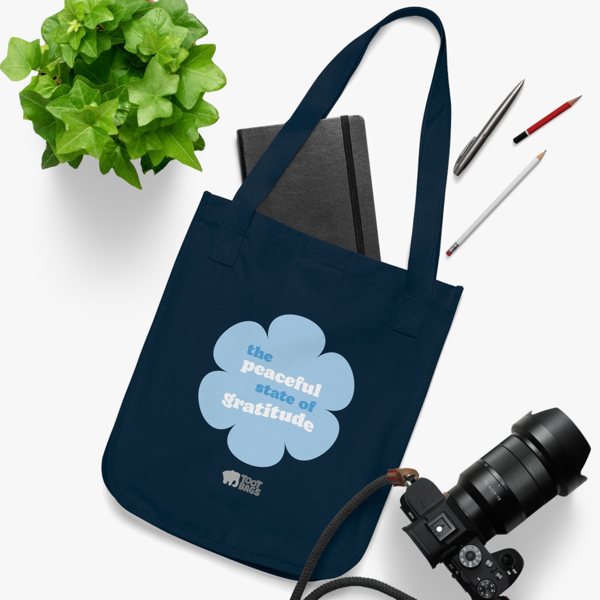 Navy blue canvas tote bag with a printed design featuring a blue flower and ‘the grateful state of gratitude’ text design placed on a white surface with stationery items, a plant and a camera.