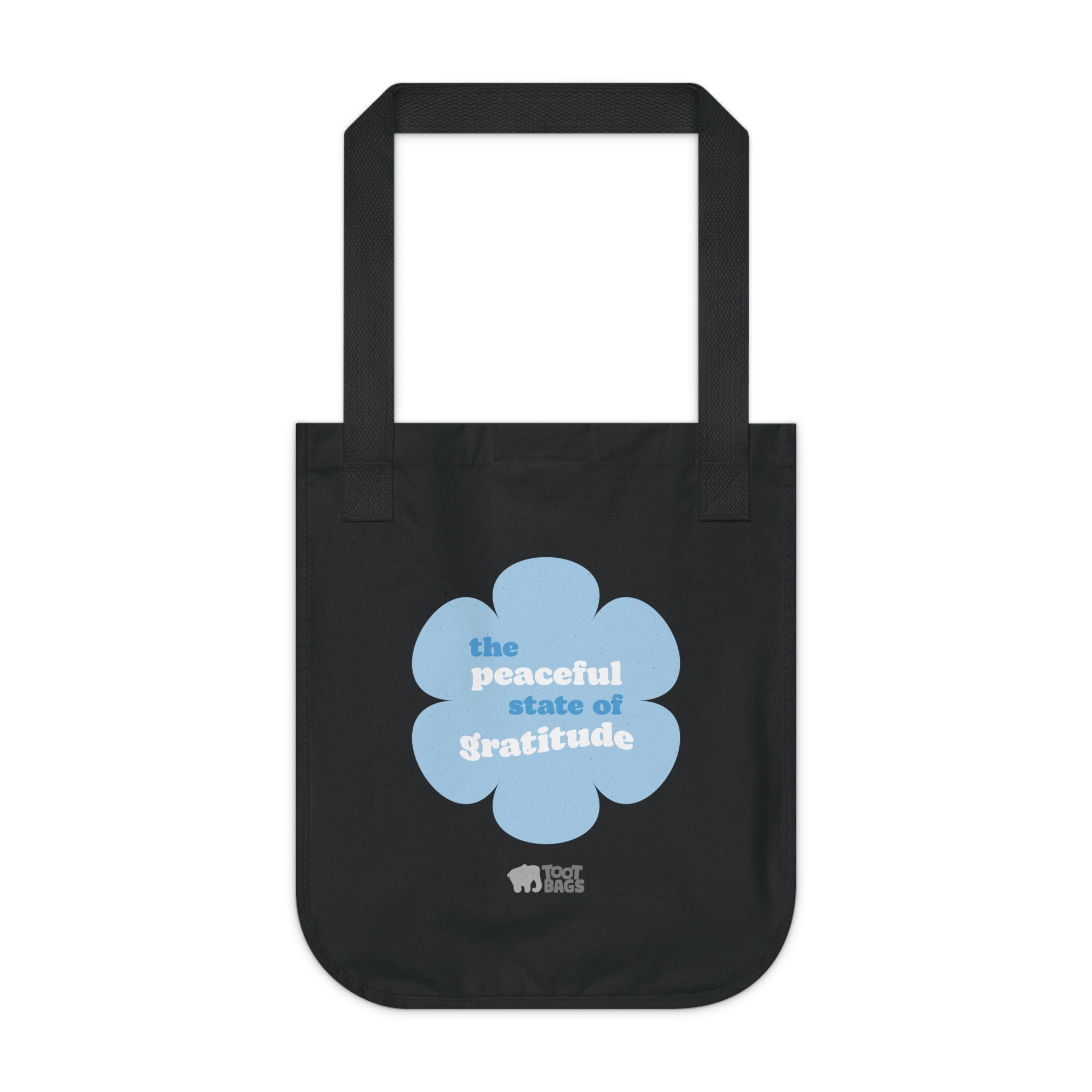 Black canvas tote bag with a printed design featuring a blue flower and ‘the peaceful state of gratitude’ text design on a white background.