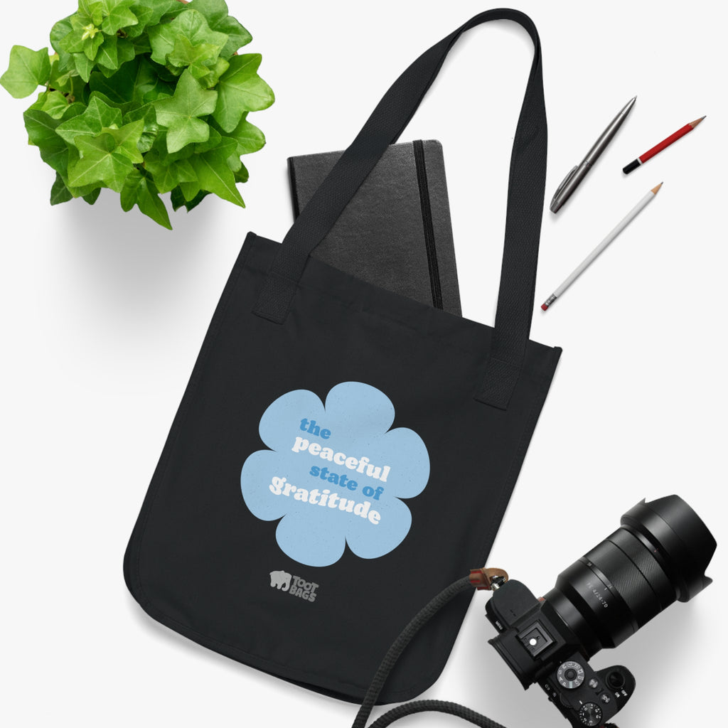 Black canvas tote bag with a printed design featuring a blue flower and ‘the grateful state of gratitude’ text design placed on a white surface with stationery items, a plant and a camera.